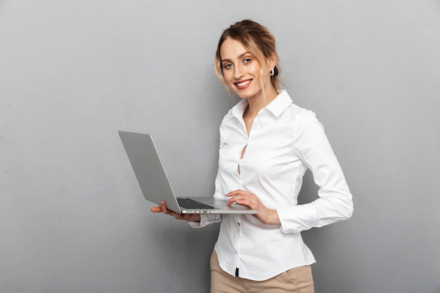 Foto di donna intelligente in abbigliamento formale in piedi e tenendo il computer portatile in ufficio, isolato