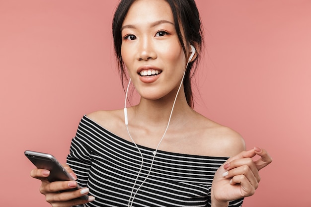 Photo of smiling asian woman dressed in basic wear holding smartphone and listening to music with earphones isolated over red wall