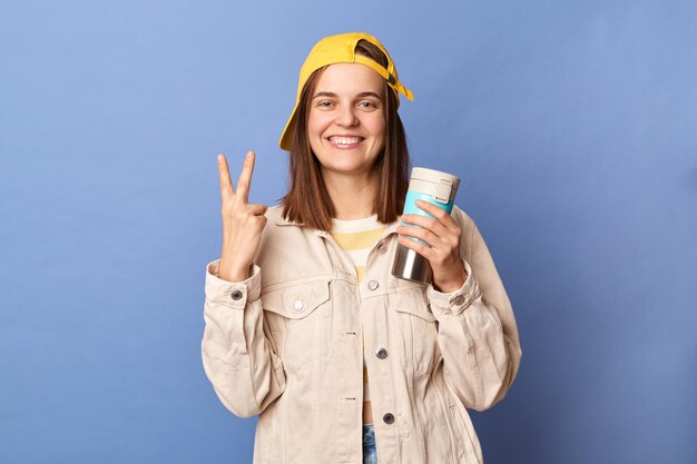 Photo of smiling delighted brown haired teenager girl wearing baseball cap and jacket drinking coffee from thermos showing victory gesture v sign posing isolated over blue background