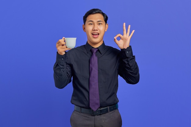 Photo of smiling handsome businessman holding cup and showing ok sign on purple background