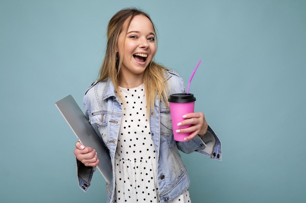 Photo of smiling happy young blonde woman wearingdenim jacket isolated over blue background carrying