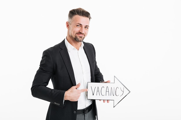 Photo of smiling successful man wearing black jacket gesturing on speech arrow pointer with word vacancy in hand, isolated over white