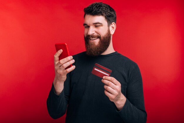 Photo of smiling young man with beard looking at smartphone and holding credit card