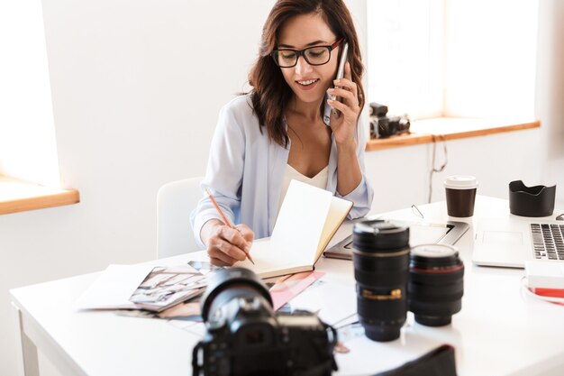 Photo of a smiling young woman photographer talking by mobile phone using laptop computer writing notes.