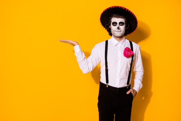 Photo of spooky ghost guy hold empty space propose interesting rare festival item hand pocket wear white shirt death costume sugar skull suspenders isolated yellow color background