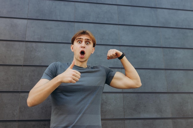 Photo of a strong athlete standing isolated over gray wall background Looking at camera pointing thumb up excited cheerful guy