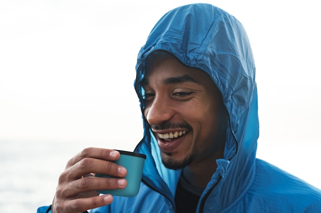 Photo of a strong handsome young african sports man outdoors at the beach sea drinking hot tea.