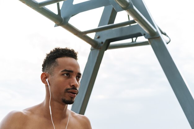 Photo of a strong handsome young african sports man outdoors at the beach sea walking make exercises listening music with earphones.