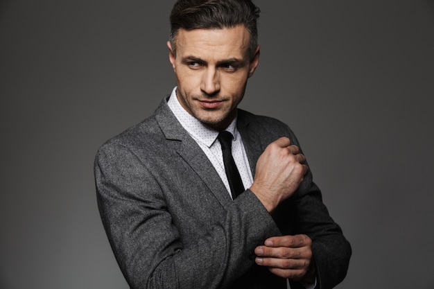 Photo of stylish man wearing business suit looking aside, while fasten cufflink or button on sleeve of jacket, isolated over gray wall