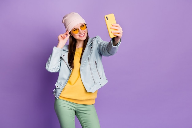 Photo of stylish pretty nice lady hold telephone smiling make selfies followers popular blogger wear sun specs casual hat blue jacket pullover green pants isolated purple color background