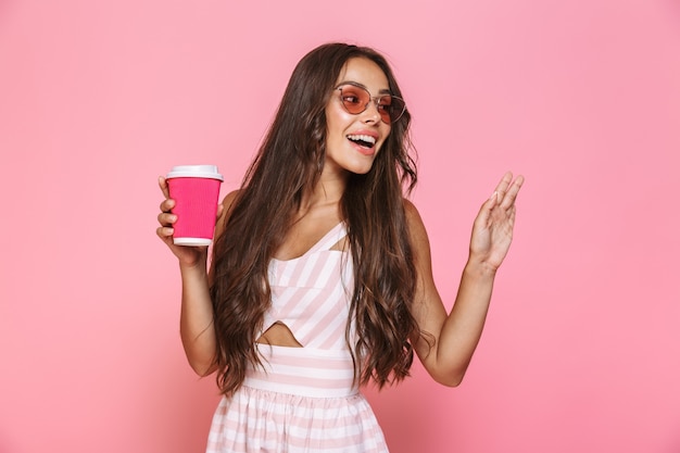 Фотография стильной женщины 20-х годов в солнцезащитных очках, смеющейся и держащей бумажный стаканчик, изолированной над розовой стеной