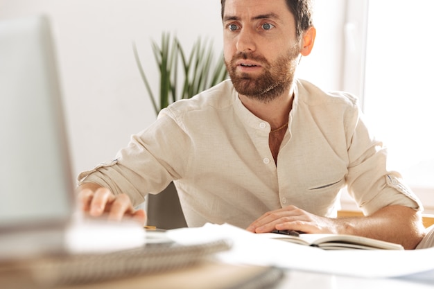 Photo of successful businessman 30s wearing white shirt using laptop, while working in bright office