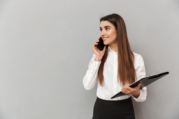 Фотография успешной женщины 20 лет в белой рубашке и черной юбке, держащей буфер обмена с файлами в офисе и говорящей по мобильному телефону, изолированной над серой стеной