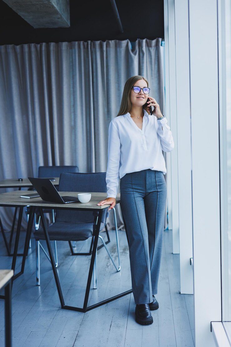 Photo photo of a successful young woman in a shirt smiling and working on a laptop while talking on the phone in a modern office with large windows remote work with coffee