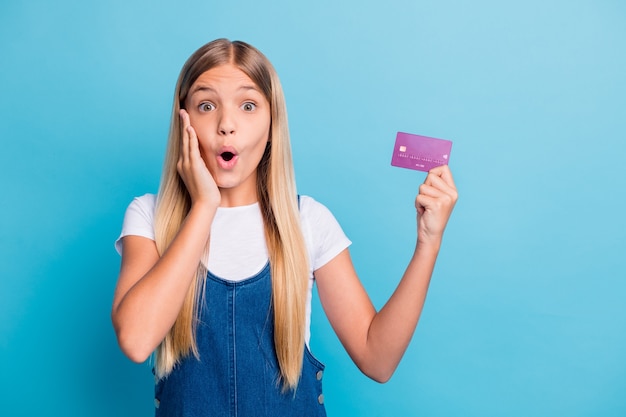 Foto foto di una ragazza adolescente sorpresa e allegra con i capelli biondi in possesso di una carta di credito che indossa abiti casual isolati su uno sfondo di colore blu pastello