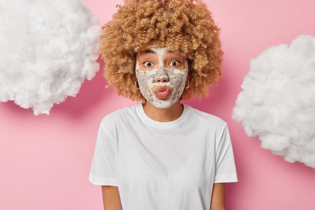 Photo of surprised young woman keeps lips rounded applies\
nourishing clay mask for reducing fine lines undergoes skin care\
beauty procedures dressed in t shirt isolated over pink\
background