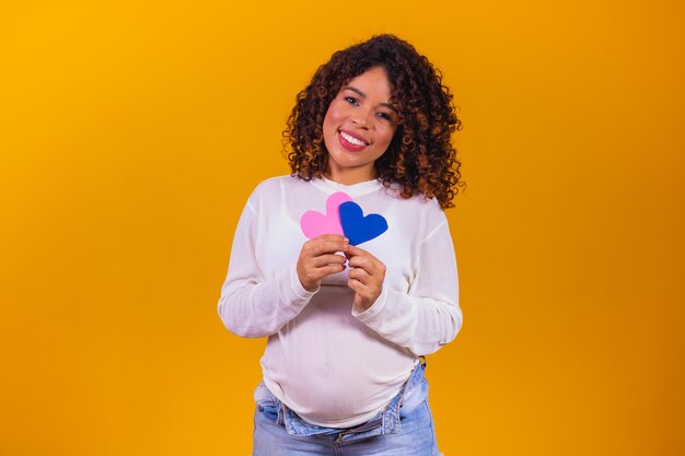 子供がピンクの青いハートの孤立した黄色の背景を保持している腹を指す白い服を着ることを期待している甘い女性の写真