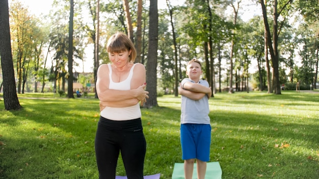 Photo of teenage boy doing yoga with his mother on grass park. Family doing fitness and stretching exercises together at forest