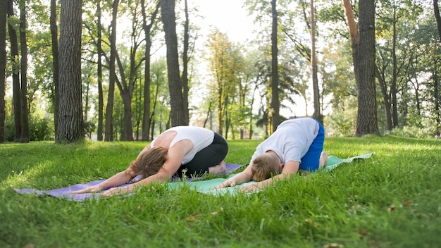 Photo of teenage boy doing yoga with his mother on grass park. Family doing fitness and stretching exercises together at forest