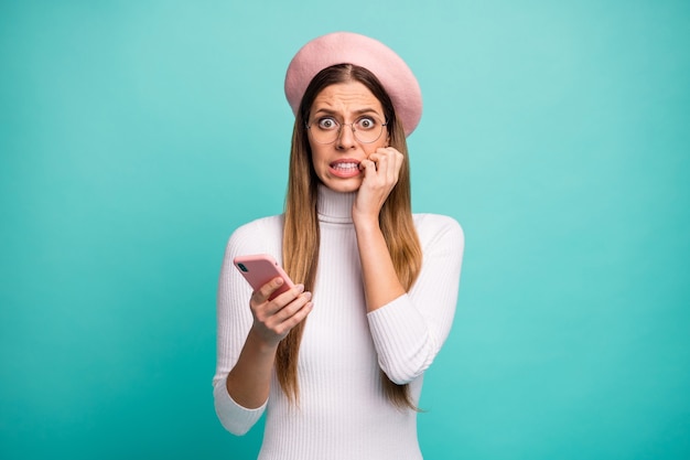 Photo of terrified freelancer lady hold telephone read post negative comments biting fingers wear specs modern pink beret cap white turtleneck isolated bright teal color background