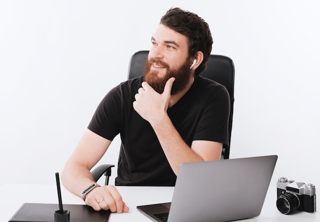 Foto di pensare ragazzo barbuto, giorno lavorativo, spazio ufficio con molti gadget