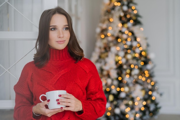 Photo of thoughtful female model wears red sweater drinks tea\
has dark hair contemplates about something stands in spaciouss room\
near decorated new year tree with lights holiday concept