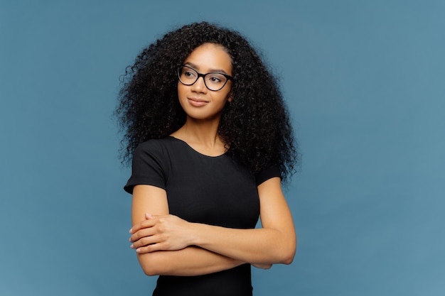 Photo of thoughtful satisfied Afro woman keeps hands crossed over chest focused aside wears transparent glasses casual black t shirt isolated over blue background Human facial expressions concept