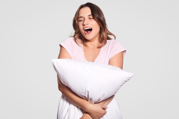 Photo of tired attractive female model yawns, opens mouth widely, dressed in casual clothes, carries pillow, needs to have good rest, isolated over white wall. Woman hugs cushion indoor