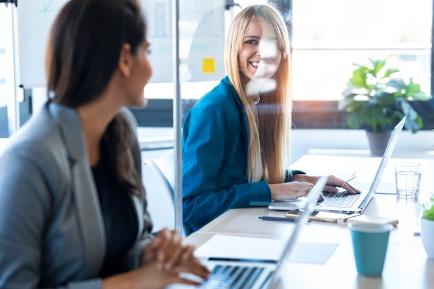 Photo of two business women looking at each other as they work with laptops at the divided desk in the coworking space. Concept of social distancing.