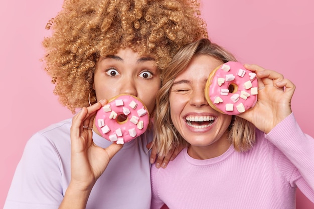 Photo of two female best friends hold delicious doughnuts with marshmallow have sweet tooth enjoy eating tasty unhealthy food dressed casually isolated over pink background Harmful nutrition