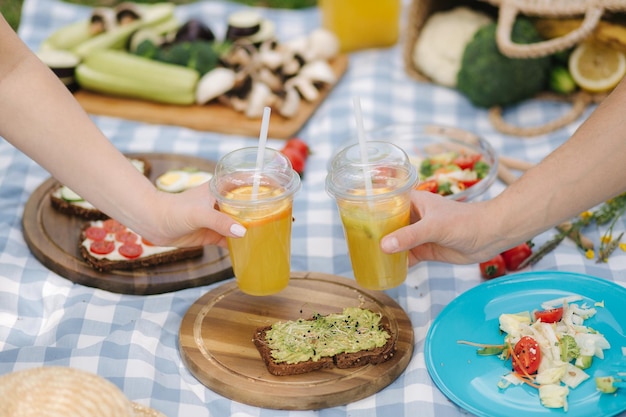 Photo of two hand hold orange lemonade in front of vegan picnic outdoors Healthy food concept