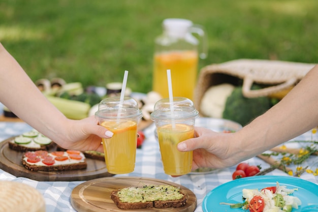 Photo of two hand hold orange lemonade in front of vegan picnic outdoors Healthy food concept