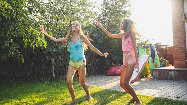 Photo of two happy laughing sisters in wet clothes dancing under water droplets from garden hose at garden. Family playing and having fun outdoors at summer