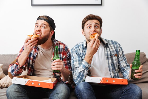 Photo of two manly men in casual shirts eating pizza and drinking beer, while watching football game at home