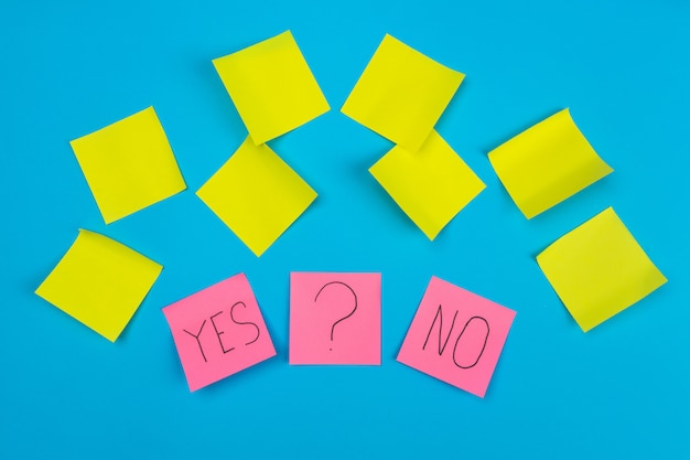 Photo photo two pink stickers with the phrase yes or no and a sticker with a question mark on a blue  with yellow self-adhesive stickers.  of office stationery.