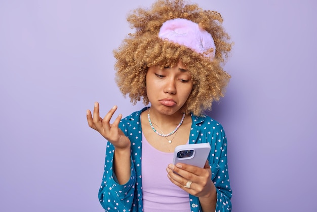 Photo of unhappy frustrated woman looks sadly at smartphone\
screen finds out bad news wears soft sleepmask and blue pajama\
isolated ovr purple background bad emotions and technology\
concept