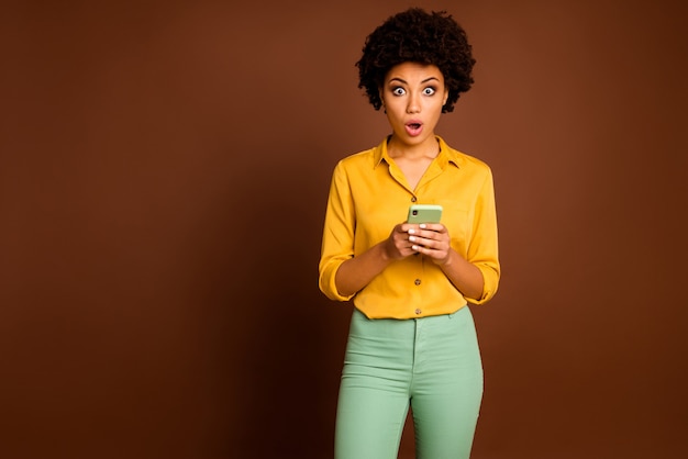 Photo of upset dark skin wavy lady holding telephone app freelancer open mouth read awful negative comments wear yellow shirt green pants isolated brown color