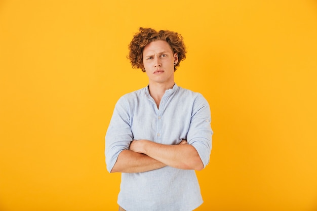 Photo of upset serious man  with curly hair standing with arms folded, isolated over yellow background