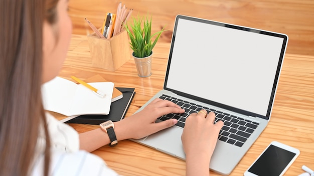 Photo views from back of young woman while using/typing on white blank screen laptop including pencil holder, black blank screen smartphone, potted plant, pencil, notes, earphone on wooden desk.