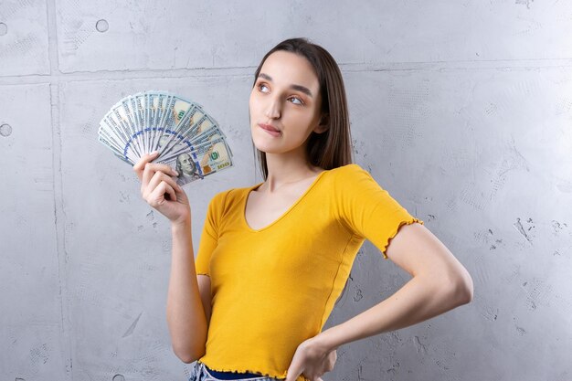 Photo of a wealthy woman in simple clothes holding a fan of dollar money isolated against a concrete wall background