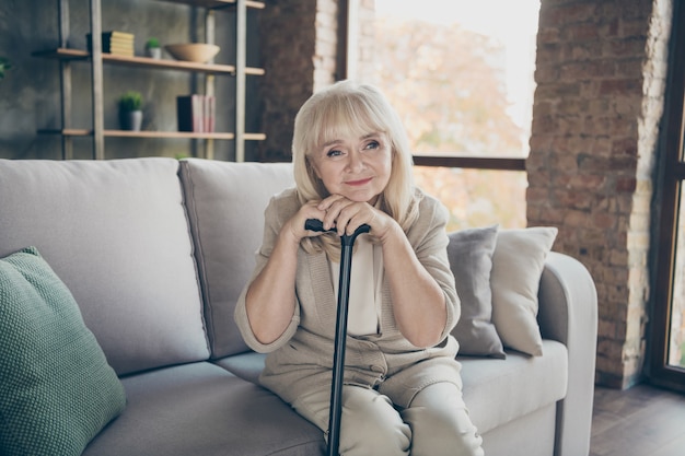 Photo of white haired aged grandma holding walking cane remembering youth light sadness romantic memories missing husband sitting divan sofa living room apartments indoors