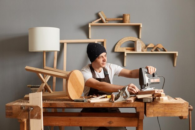 Photo of young adult man carpenter wearing white tshirt black cap and brown apron joiner being engaged in woodworking using electric jigsaw posing among joiner equipment