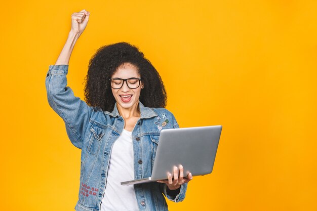 Photo of young african american cheerful business woman standing isolated over yellow background with laptop computer. looking aside make winner gesture