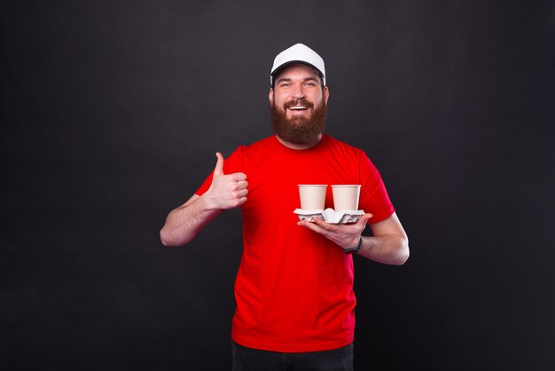 Photo of young bearded hipster man in red t-shirt holding two cups of coffee and showing thumb up