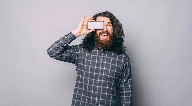 Photo of young bearded man holding smartphone over eyes