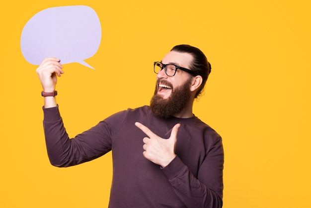 Photo of a young bearded man pointing at a speech bubble that he is holding near a yellow wall wearing glasses