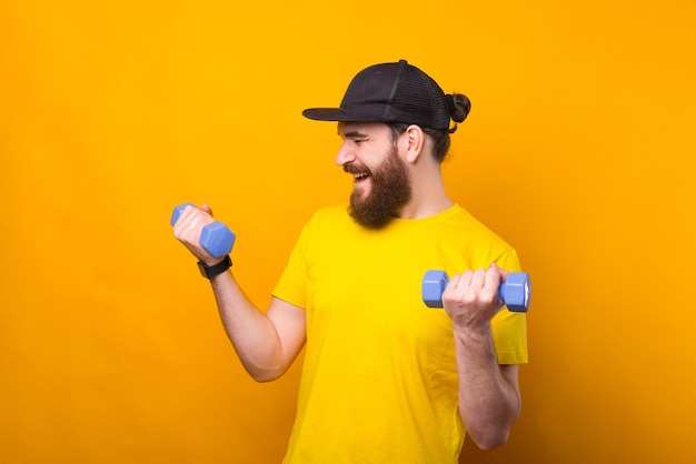 Photo of young bearded man using small dumbbells over yellow background