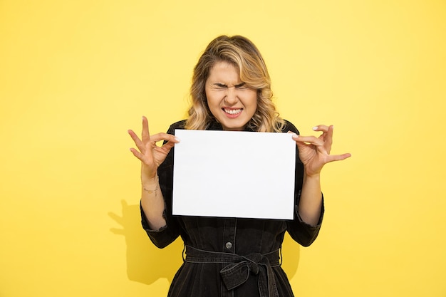 Photo photo of young beautiful blonde woman is capricious wrinkled her face, made dissatisfied expression, holding in hands clean empty white sheet of paper a4 for sign, studio shot on yellow background
