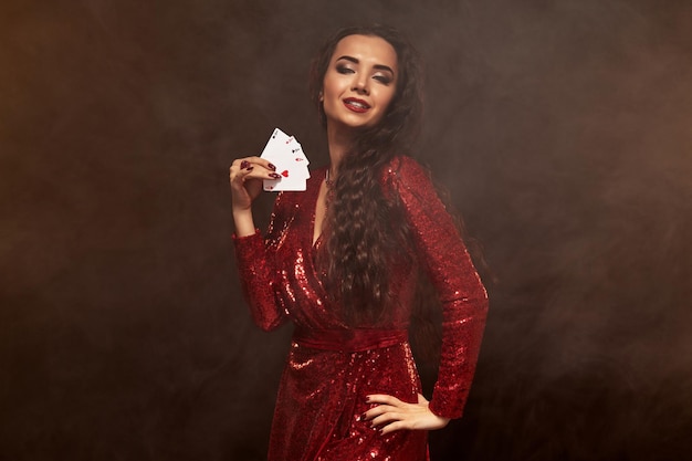 빛나는 빨간 이브닝 드레스를 입은 젊은 백인 브루네트 여성의 사진은 한 손에 카드를 들고 4개의 에이스를 보여줍니다. 갈색 배경, 카지노, 도박 산업