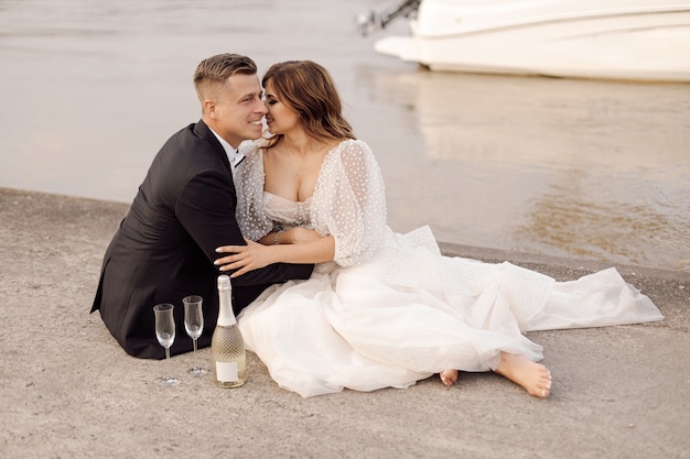 Photo of young beautiful couple, bride and groom in wedding dress are sitting on bank of wide river with glasses of wine and champagne. wedding day, happy moments, kissing .